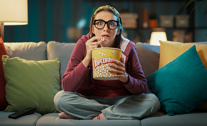 Woman sitting on her couch, eating popcorn and watching scary movies streaming from Access Video On Demand