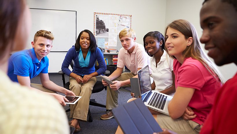 High school students having a discussion in a circle with laptops and tablets